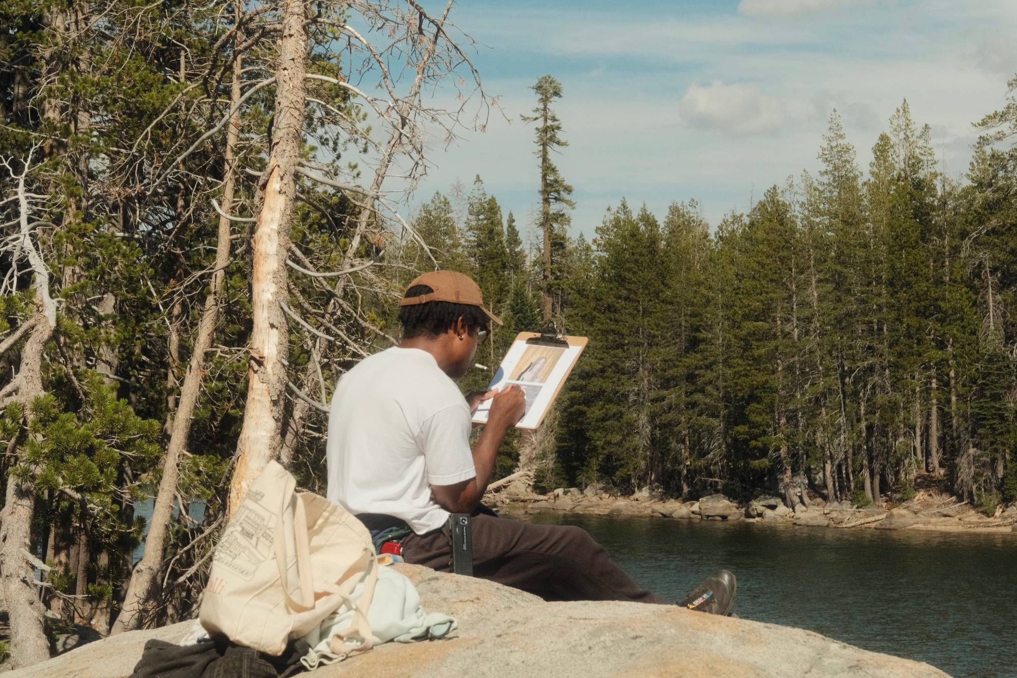 Azeem sitting on a granite boulder by a lake, coloring the Terrified cover on a clipboard